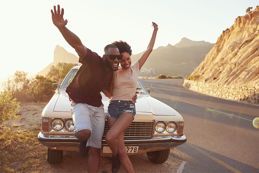 Insurance Quote - Portrait of a Smiling Young Couple Standing in Front of Their Car Parked Alongside the Road During a Road Trip on a Sunny Summer Day