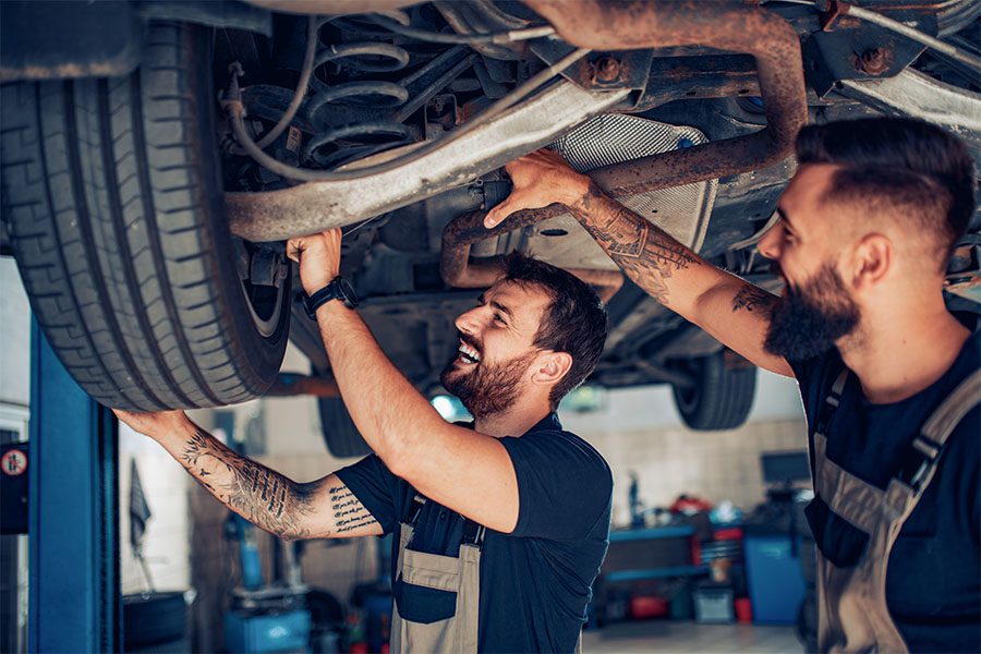 Specialized Business Insurance - Portrait of Two Cheerful Young Male Mechanics Working Underneath a Car in a Garage Repair Shop